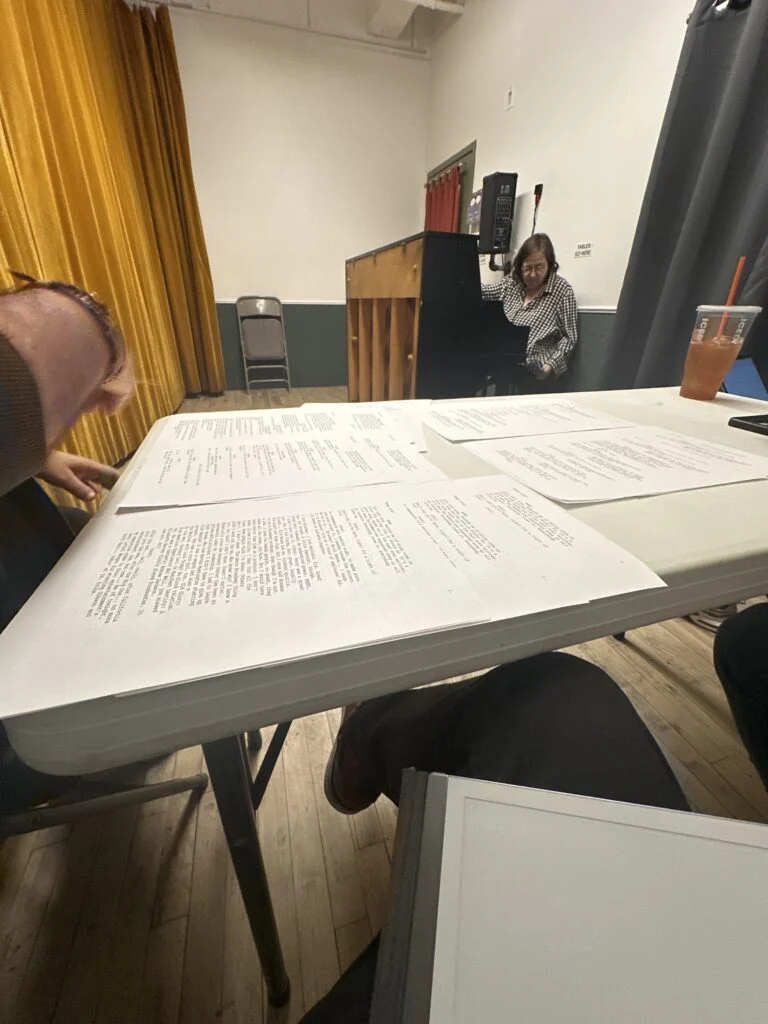 An older woman in a checkered shirt leans by a podium as scripts cover a table where another person's arm rests, in a casual rehearsal space.