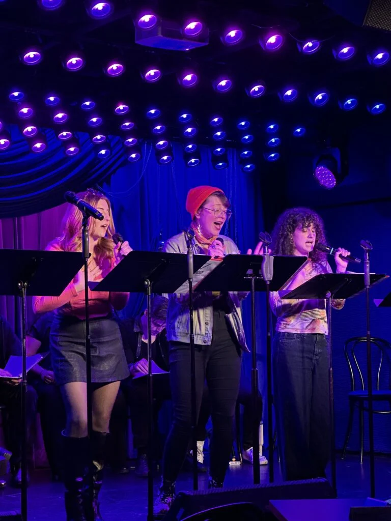 Three young adult women sing into microphones onstage under bright purple and blue lights during a vibrant performance for 'Vlad and the Impalers'.