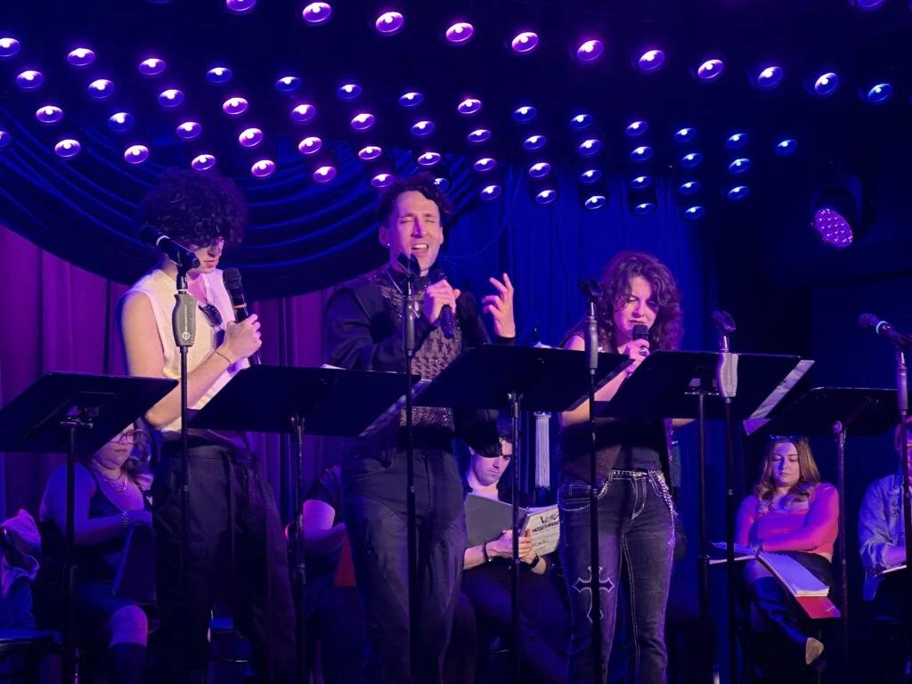 Three performers sing expressively on a dark stage bathed in vibrant purple and blue lights, creating an energetic mood.