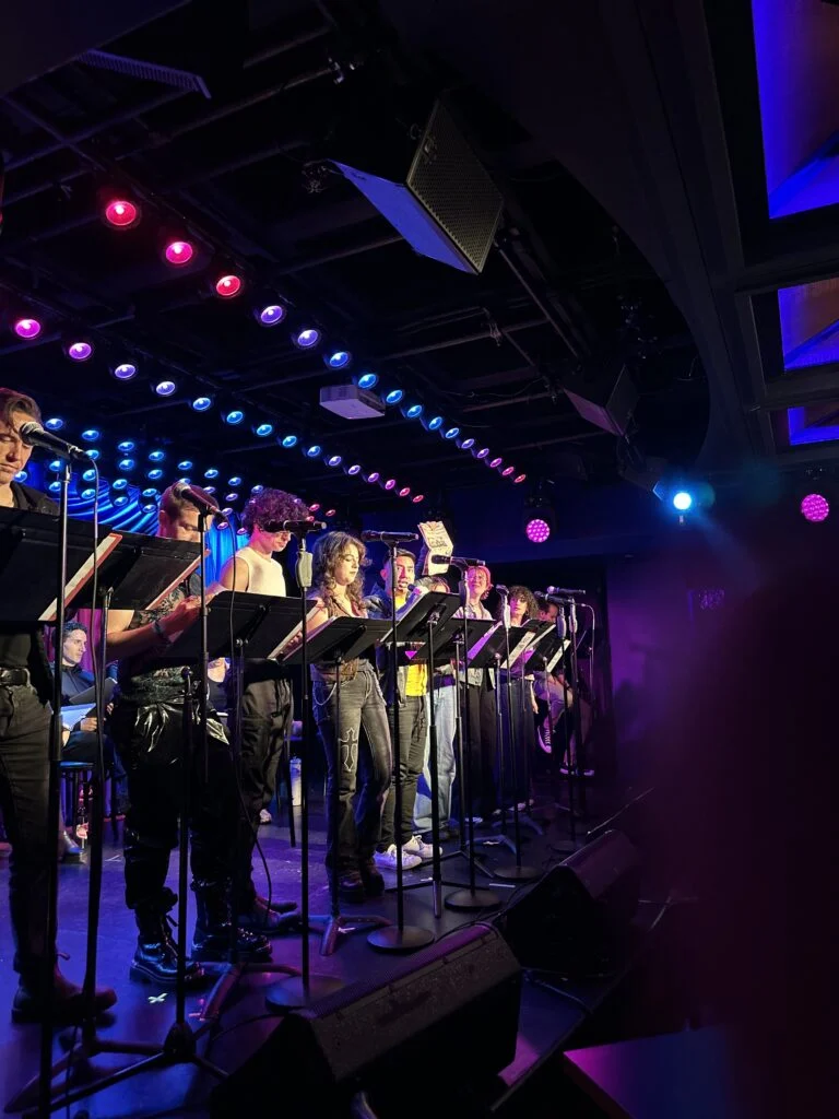 Young adult performers on a stage read from scripts at microphones under theatrical pink and blue lights for "Vlad and the Impalers".