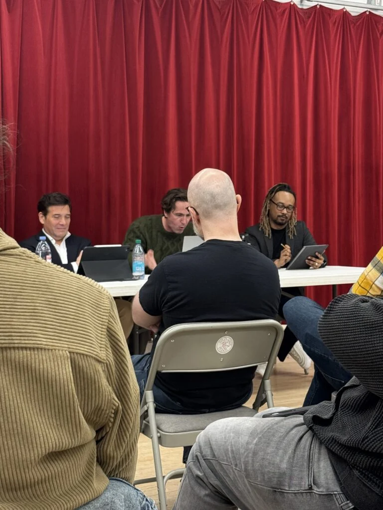 A panel discussion with a white man in a suit, a white man in a green sweater, and a Black man with dreadlocks and glasses, seated at a white table against red curtains, viewed from behind an audience member.
