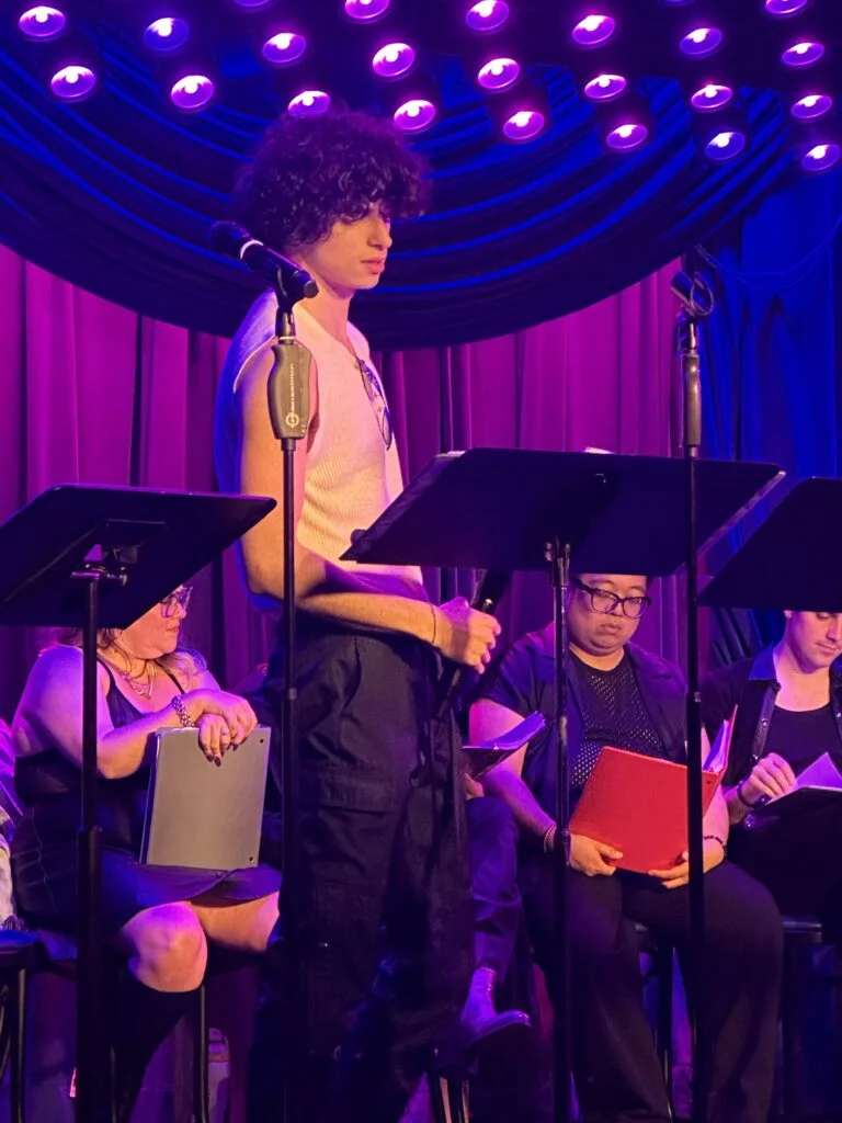 A young person with dark curly hair at a microphone on stage, flanked by seated performers, under dramatic purple and blue lights.