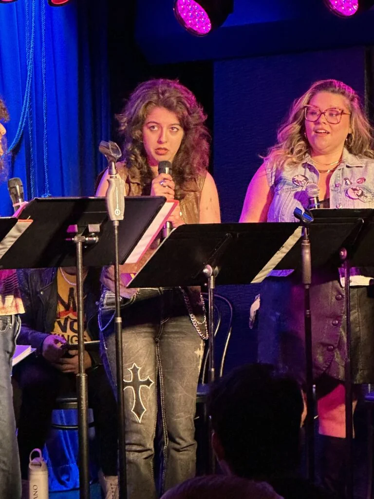 Vlad and the Impalers" performers: a woman with curly brown hair in a leather vest speaks into a mic, next to a smiling blonde woman in a denim vest on a purple-lit stage.